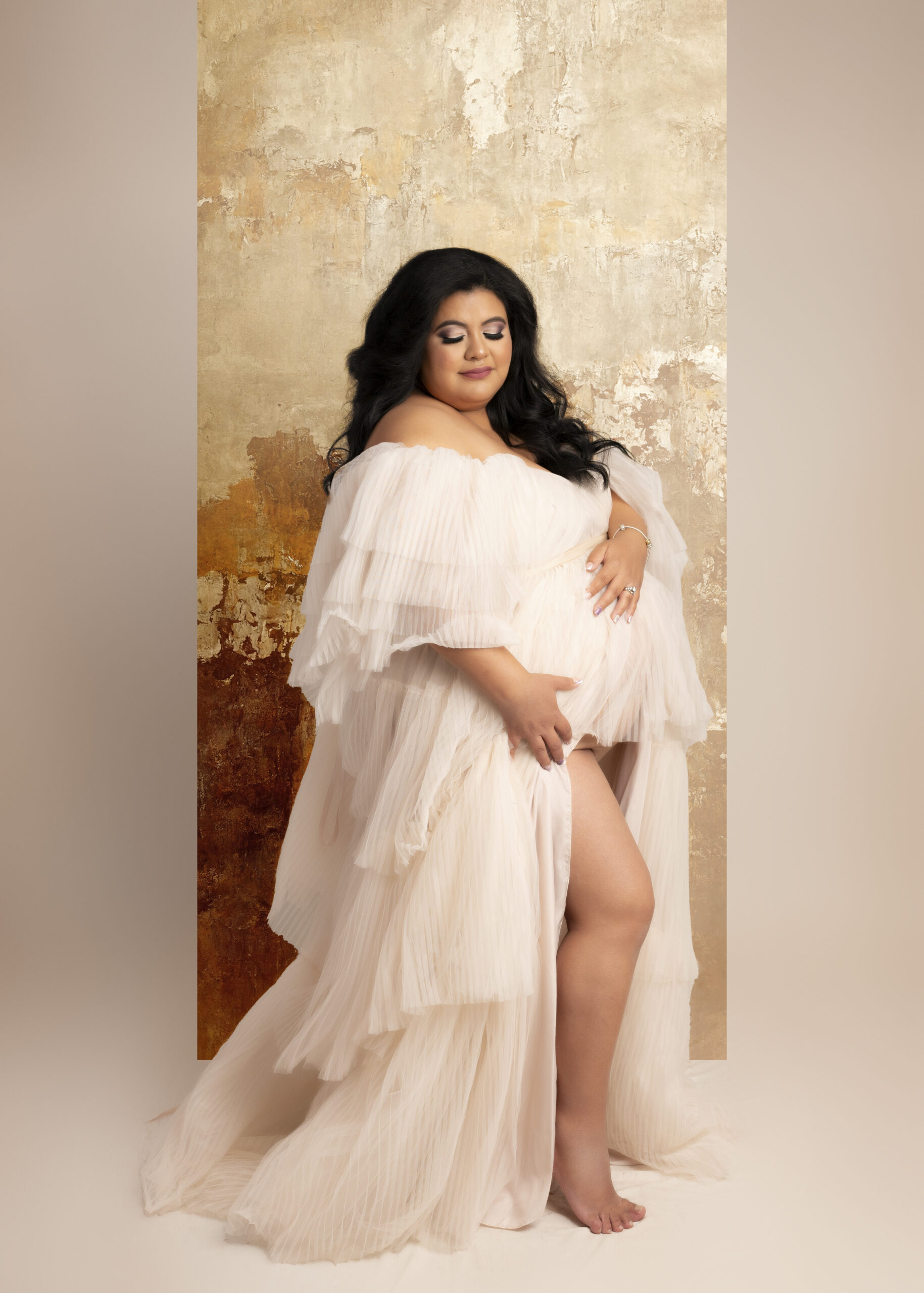 Grand rapids maternity photo session woman in long white gown with gold backdrop holding baby bump